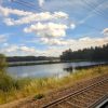 View from train window of track and blue lake and skies with trees behind.